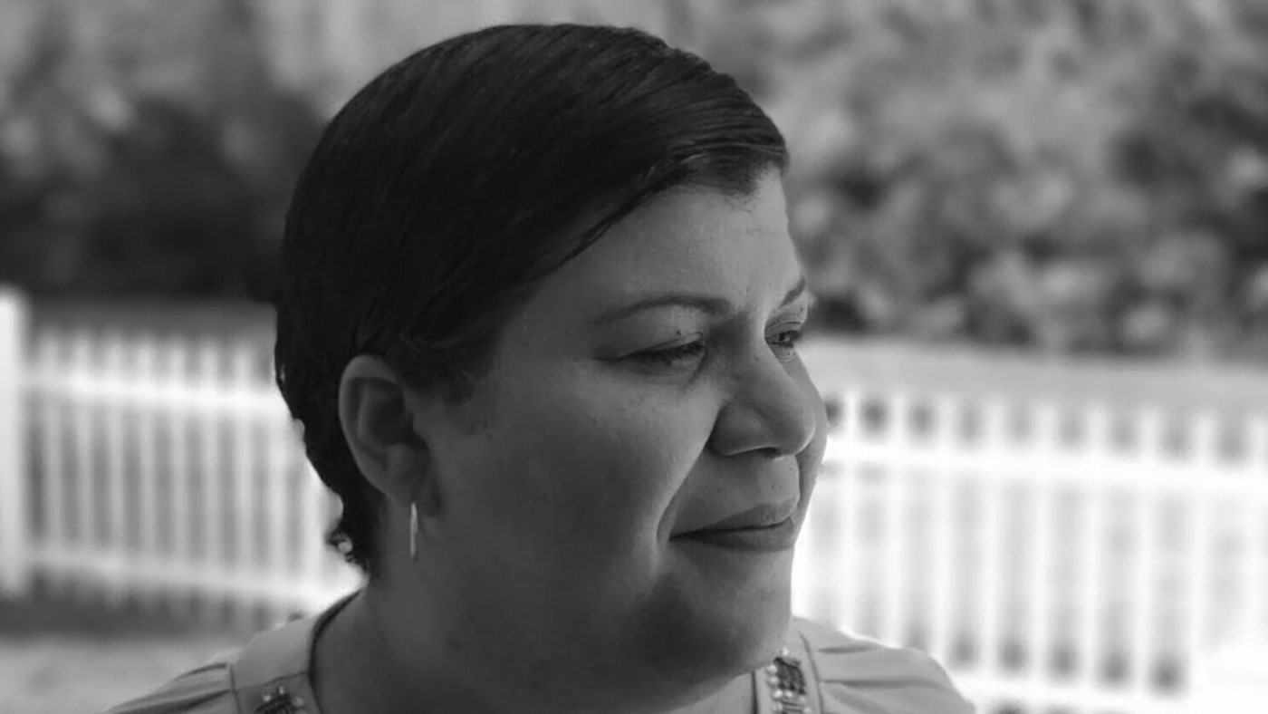 Black and white frame from the audio slide show: a woman looking off camera in front of a white picket fence